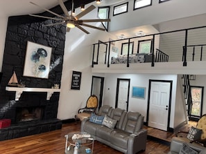 A spacious living area with a fireplace, a high ceiling fan, a staircase to an upper level, and a view of the outdoors.