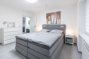A modern bedroom with a grey bed, white dresser, and a painting of trees.