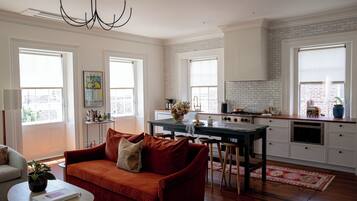 Cocina moderna con armarios blancos, una isla central y un sofá rojo en un luminoso salón con grandes ventanales.