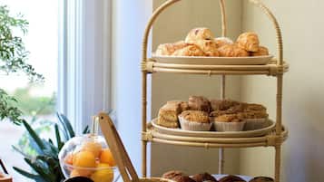 A three-tiered wicker stand with pastries and muffins, a glass bowl with oranges, and a menu sign on a wooden table by a window.