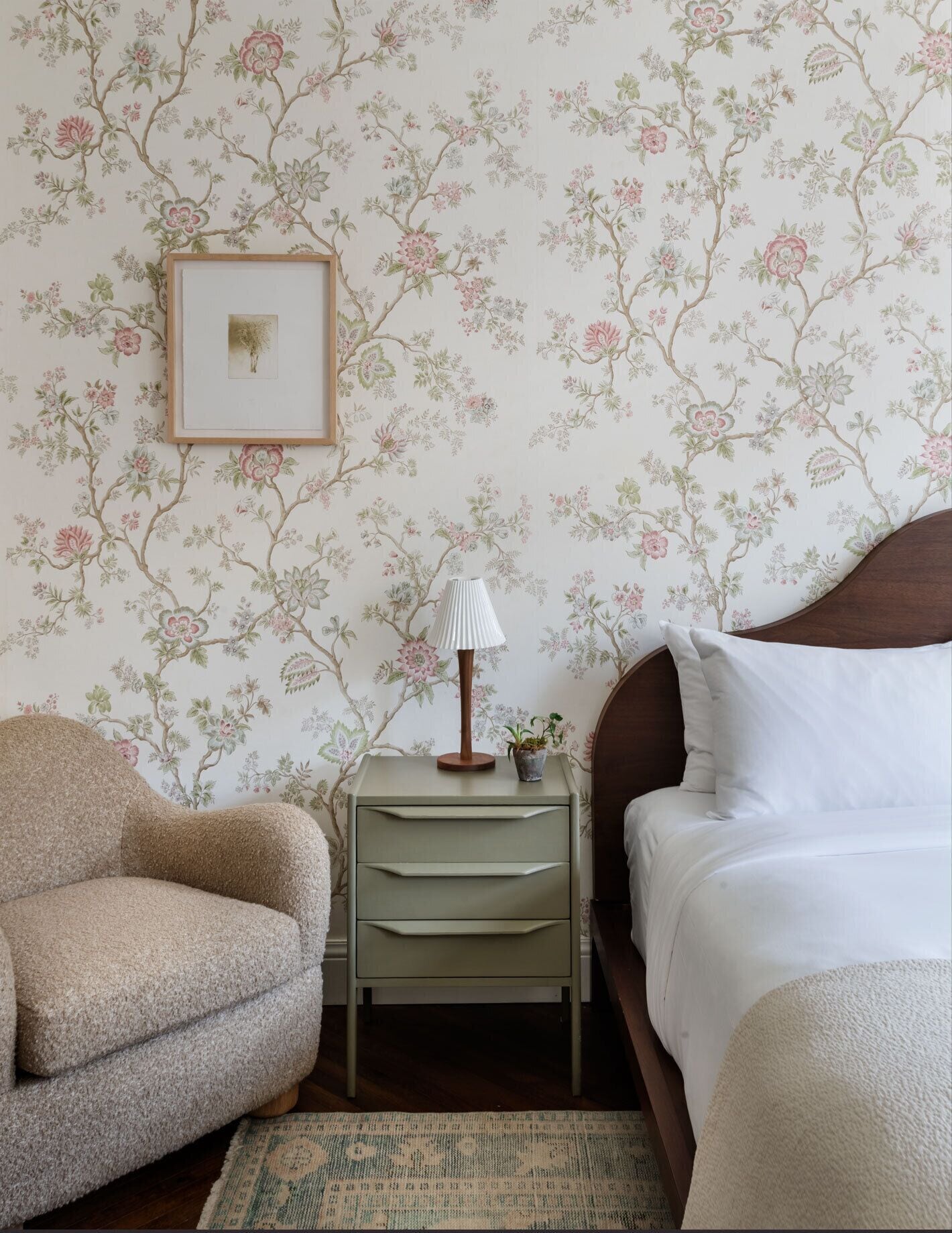 Um quarto com papel de parede floral, uma mesa de cabeceira com abajur, uma cadeira e uma cama.