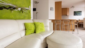 Uma sala de estar moderna com um sofá de couro branco, duas almofadas verde-claras e um banquinho redondo. Há uma parede verde com plantas decorativas e uma pequena mesa com cadeiras. Uma área de cozinha é visível ao fundo, com armários de madeira e uma mesa de jantar.