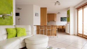 Uma sala de estar moderna com sofá branco, almofadas verdes, uma mesa de centro e uma área de cozinha com armários de madeira e uma mesa de jantar.