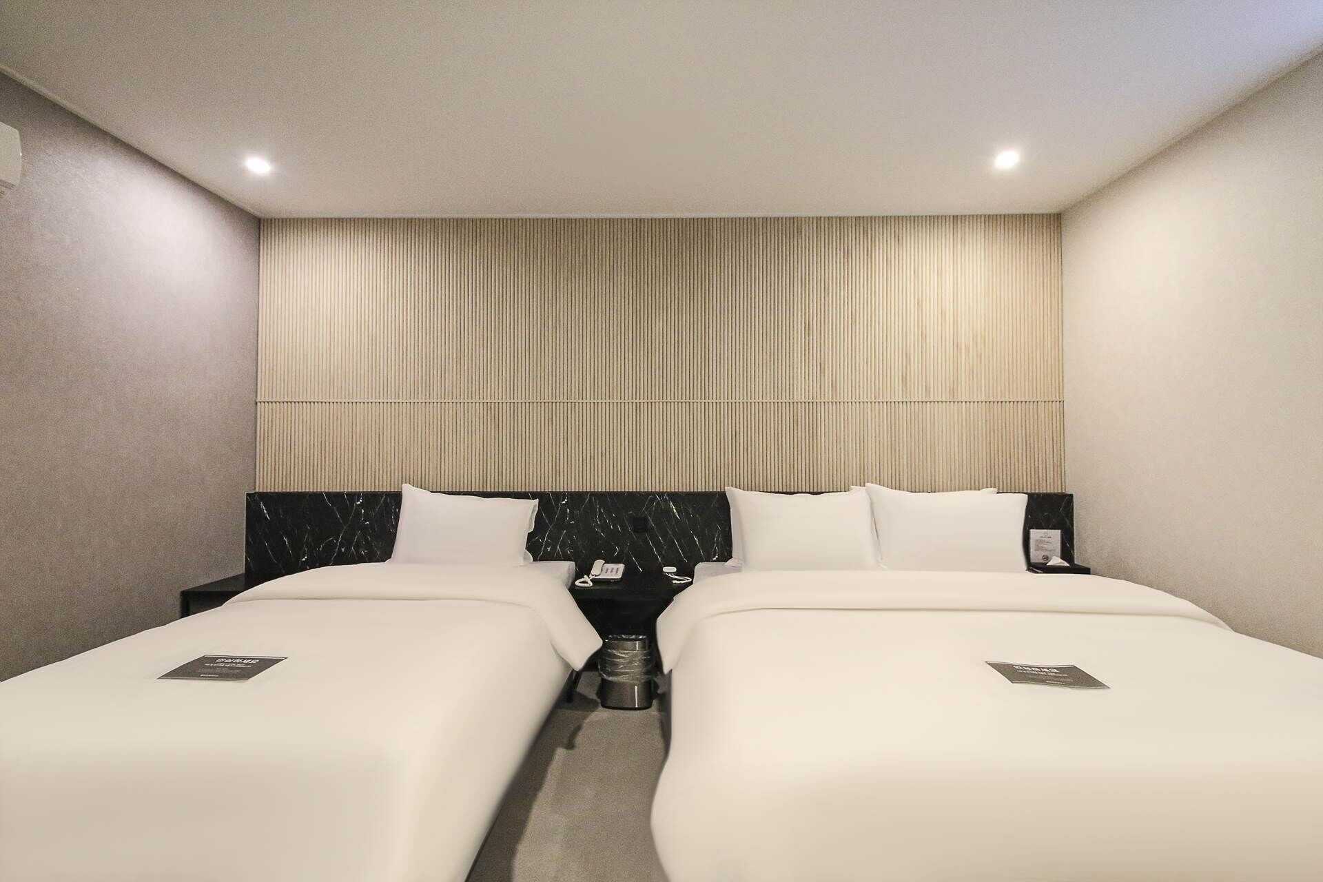 Habitación de hotel con dos camas, cabecera con líneas verticales, mesita de noche con lámpara y pequeña mesita con teléfono.
