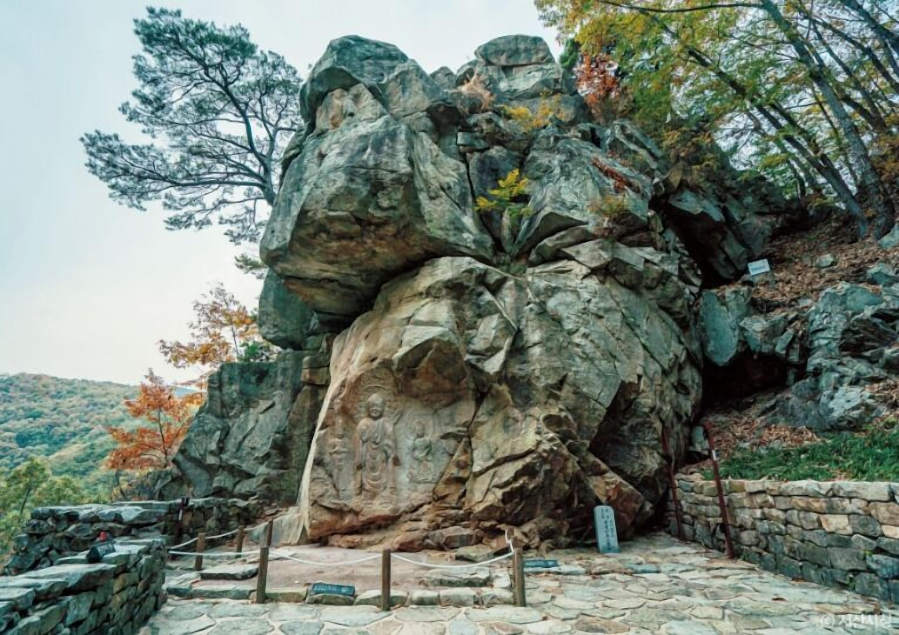 A large rock formation with a Buddha statue carved into it, surrounded by a stone pathway and a stone wall.