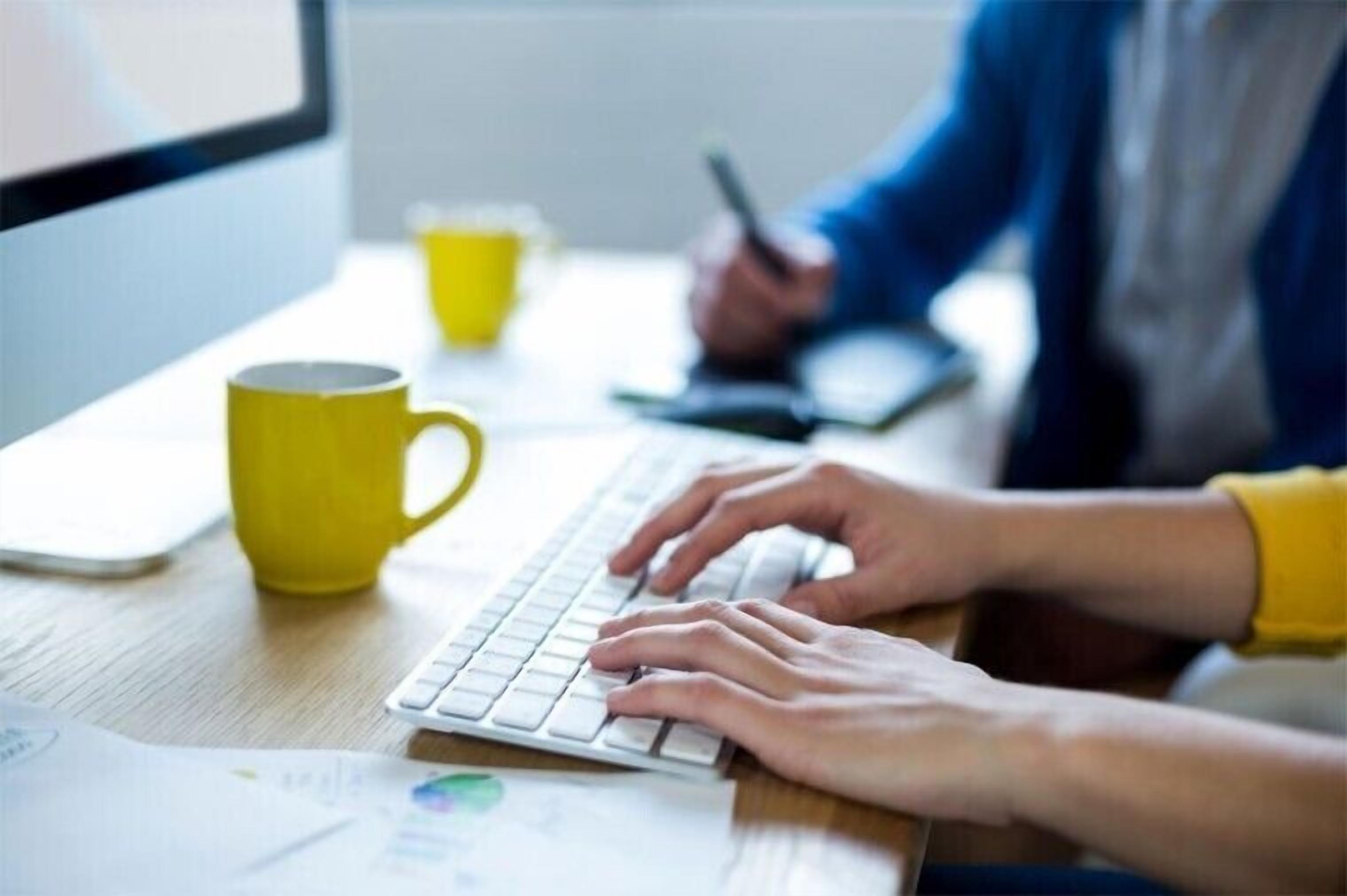 Une personne est en train de taper sur un clavier, avec une tasse jaune et des papiers sur un bureau.