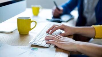 Une personne est en train de taper sur un clavier, avec une tasse jaune et des papiers sur un bureau.