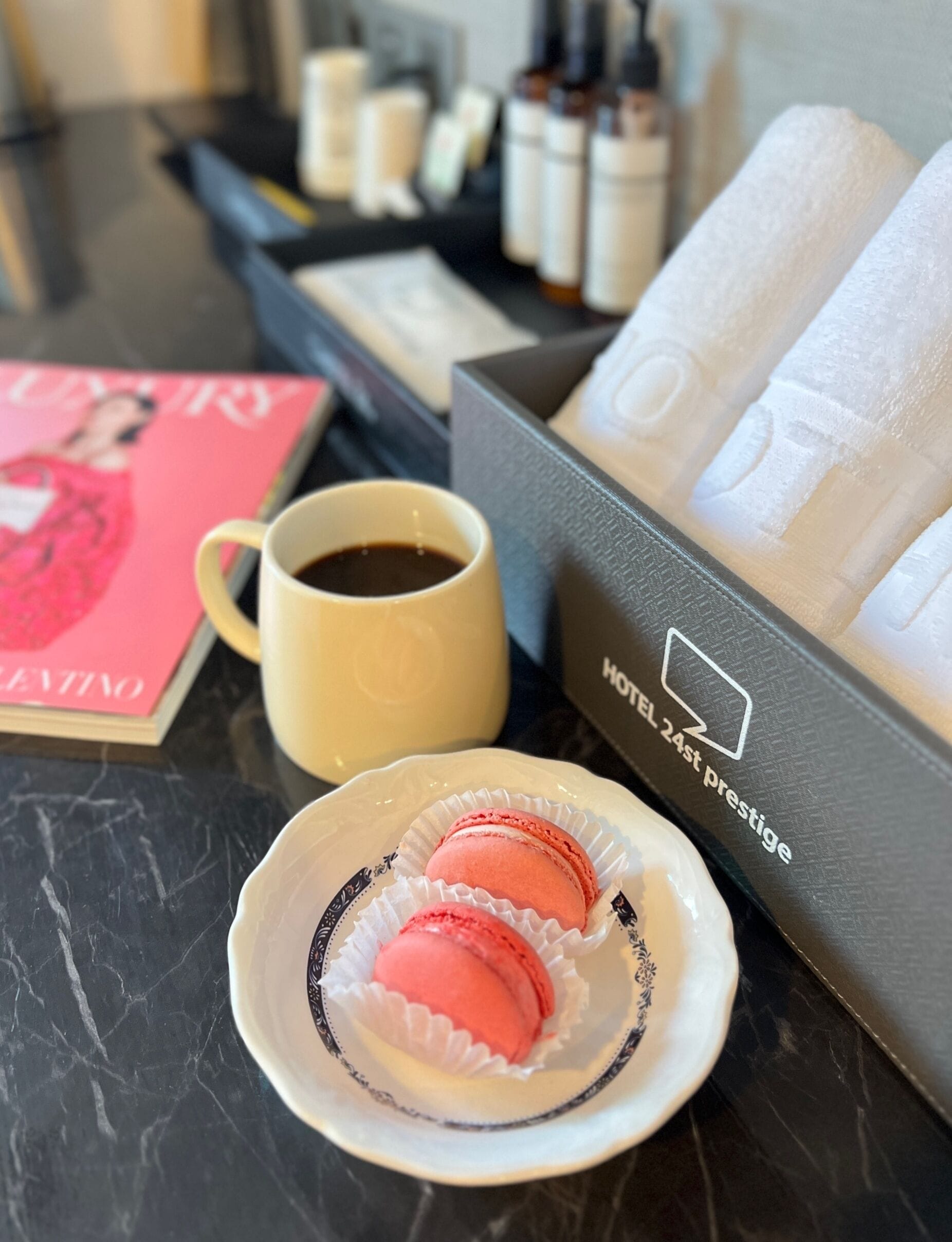 A tray with a cup of coffee, two macarons, a book, and a box labeled "HOTEL 24th prestige".