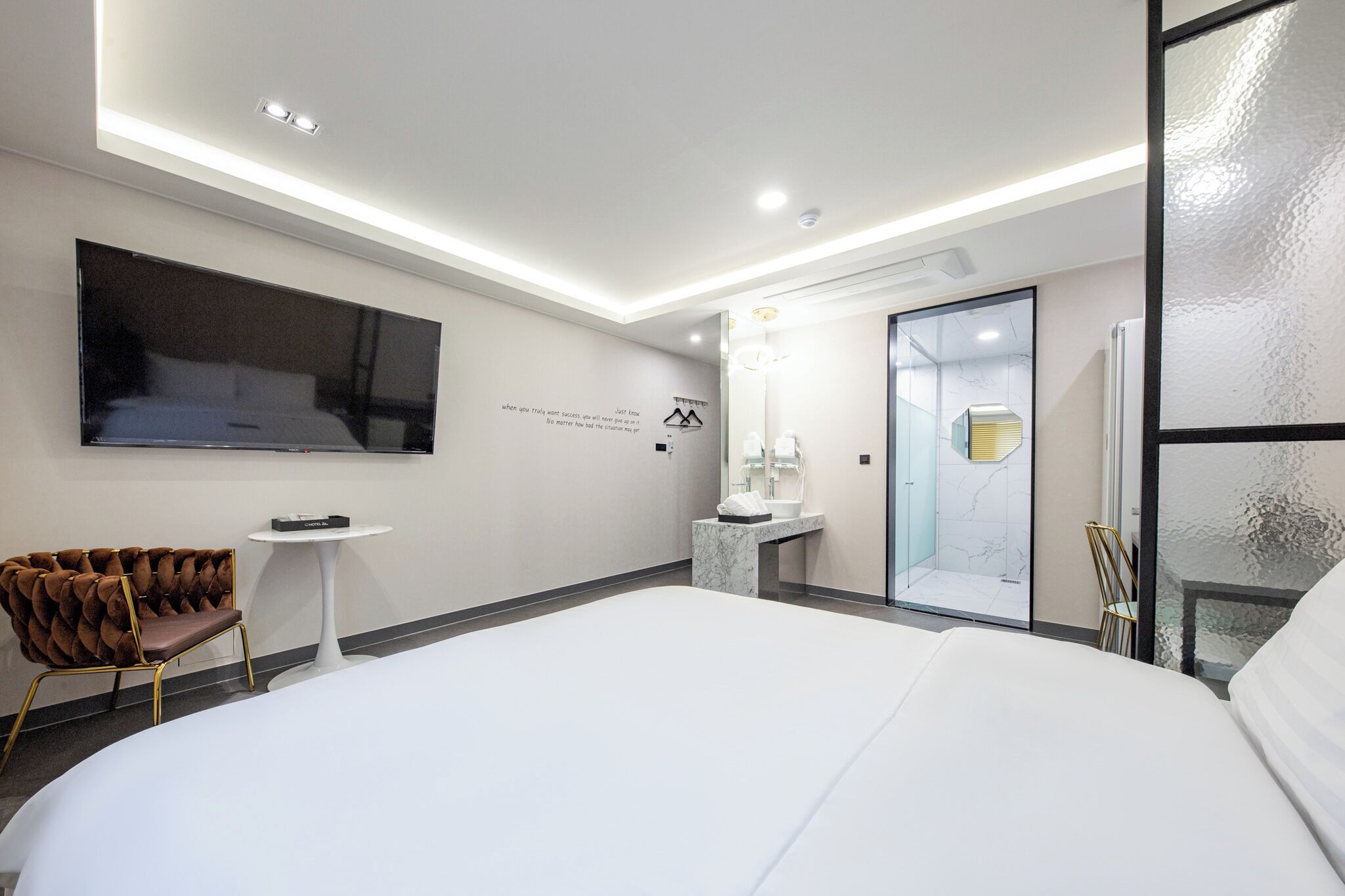 A modern hotel room with a large bed, a flat-screen TV mounted on the wall, a small round table, a tufted chair, a marble bathroom vanity, and a glass shower enclosure.