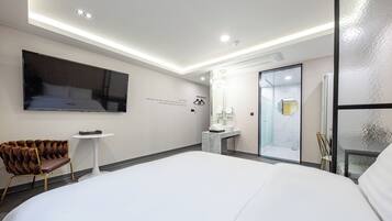 A modern hotel room with a large bed, a flat-screen TV mounted on the wall, a small round table, a tufted chair, a marble bathroom vanity, and a glass shower enclosure.