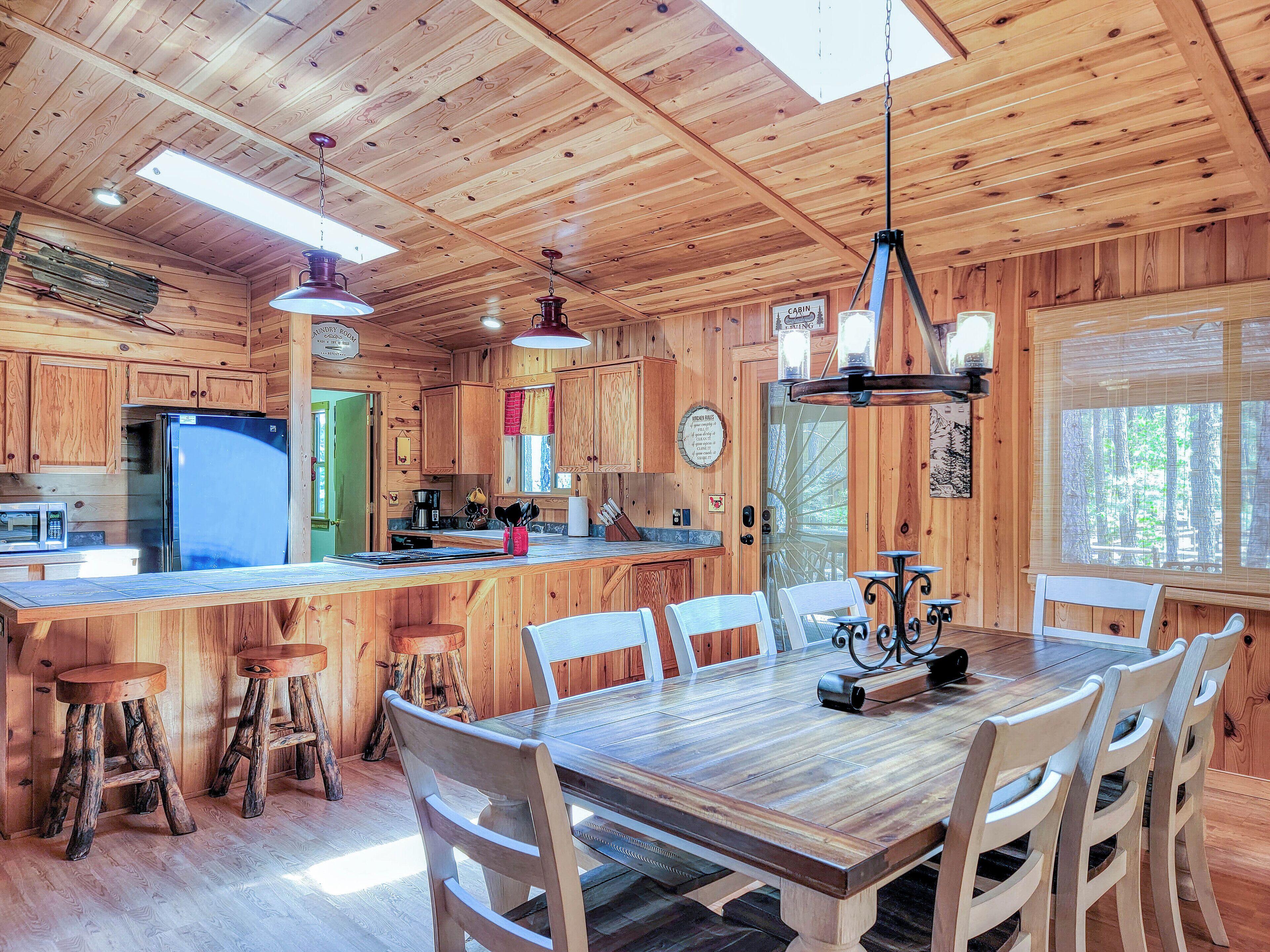 L’intérieur d’une cabane comprend une table à manger en bois, des chaises, un coin cuisine avec un réfrigérateur et une grande fenêtre.