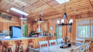 A cabin interior with a wooden dining table, chairs, a kitchen area with a refrigerator, and a large window. - White Oak Retreat in Pinetop Country Club w/Firepit/Grill/Horseshoes! (Pinetop)