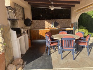 Una cocina al aire libre con pared de piedra, armarios de madera y una mesa redonda con sillas.