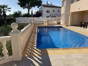 Una piscina con barandilla blanca, un suelo de baldosas y un edificio de fachada blanca al fondo.