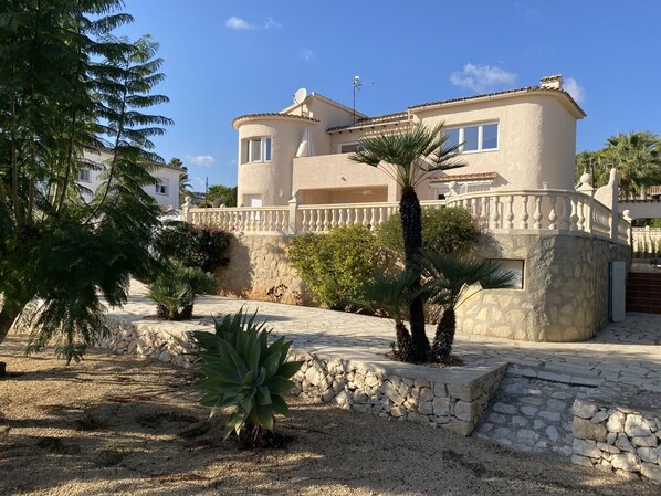 Una villa de estilo mediterráneo con fachada de piedra, un balcón y un jardín bien cuidado con palmeras y arbustos.