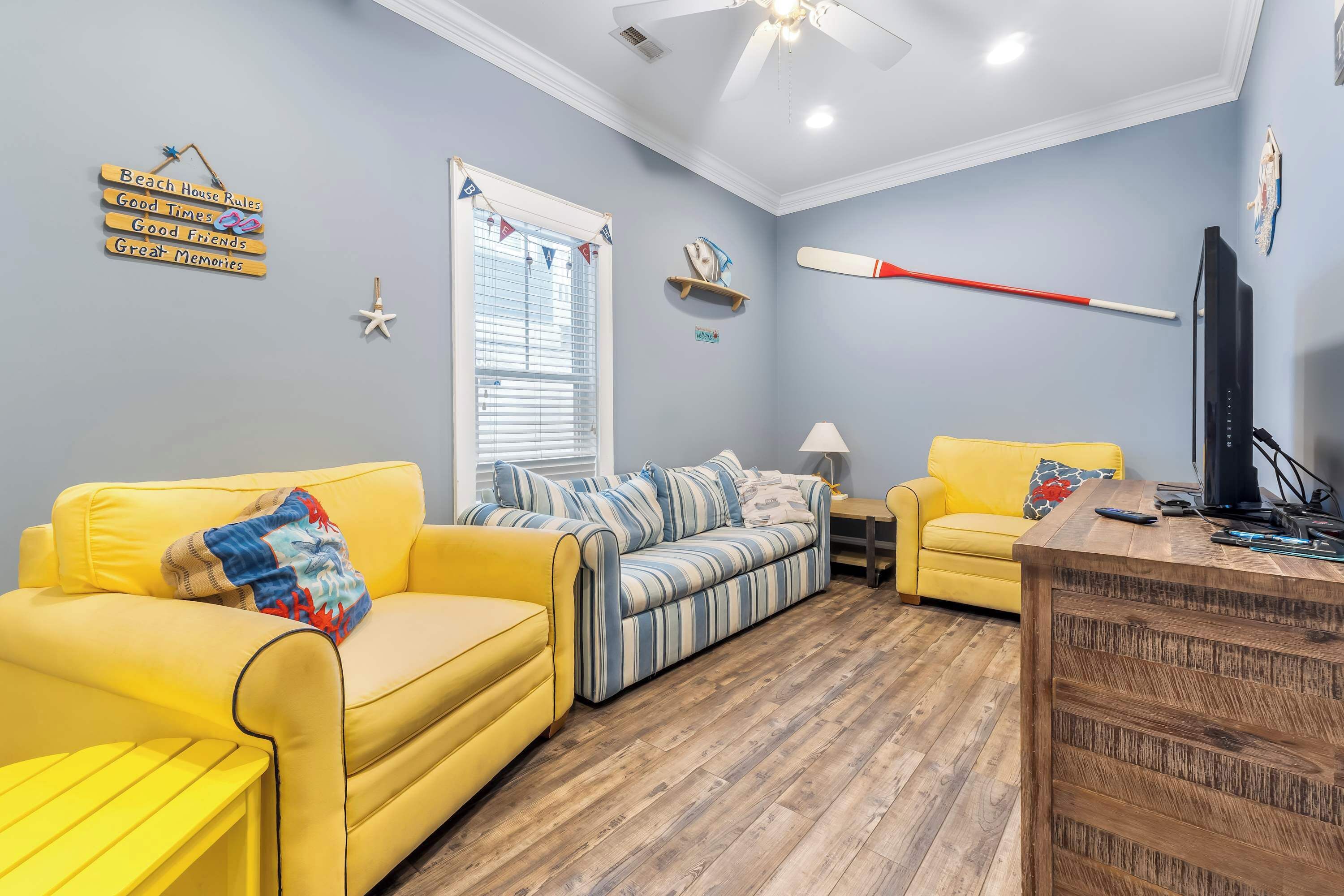 A living room with a yellow sofa, a striped sofa, a wooden coffee table, a TV, and a ceiling fan.