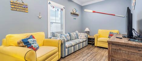 A living room with a yellow sofa, a striped sofa, a wooden coffee table, a TV, and a ceiling fan.