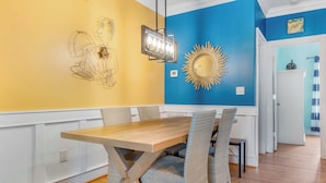 A dining area with a wooden table and wicker chairs, a wall with a sunburst mirror, and a light fixture.
