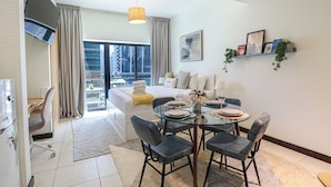 Quarto de hotel com cama, mesa de jantar posta com pratos e copos, sofá, televisão e vista da cidade por grandes janelas.