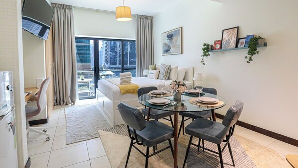 A hotel room with a bed, a dining table set with plates and glasses, a sofa, a TV, and a view of the city through large windows.