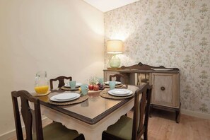 A dining area with a wooden table set for four, surrounded by chairs. A lamp is on the table, and a pitcher of orange juice is visible. - Gaya Avenue 102 - Flat w/ Balcony (Vila Nova de Gaia)