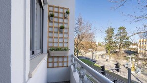 A balcony with a white railing, a wooden grid planter with plants, and a view of a street with trees and buildings. - Gaya Avenue 102 - Flat w/ Balcony (Vila Nova de Gaia)