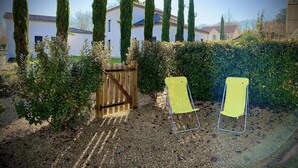 Un jardin avec une porte en bois, deux chaises jaunes et de grands cyprès.