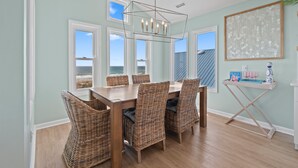 Un comedor con mesa de madera y sillas de mimbre, ventanas amplias con vista a la playa, y una lámpara de techo.