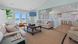 Un amplio salón con un ventanal grande con vistas al océano, una cocina con armarios blancos y un comedor con taburetes.