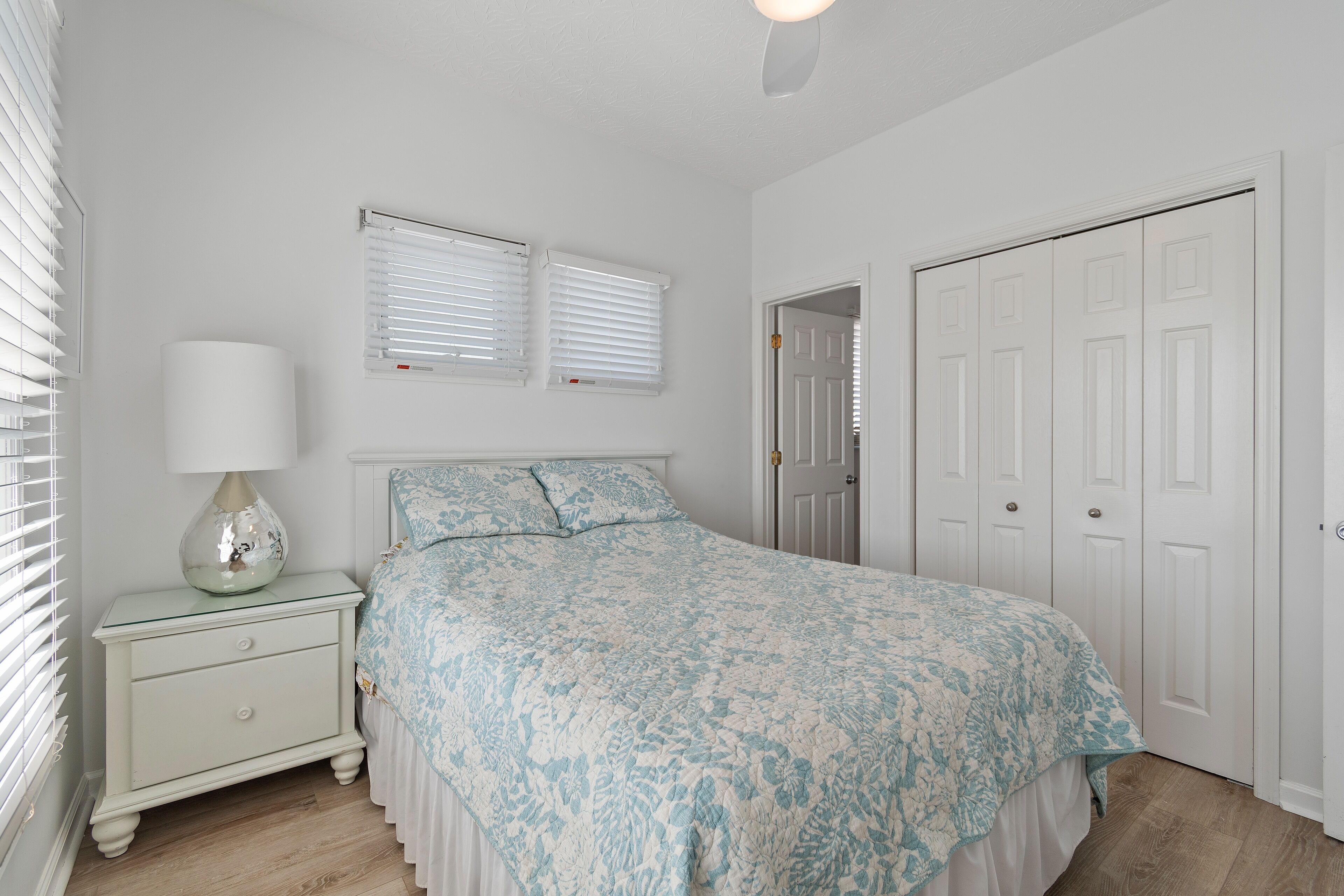A bedroom with a bed, nightstand, lamp, and two windows with blinds.