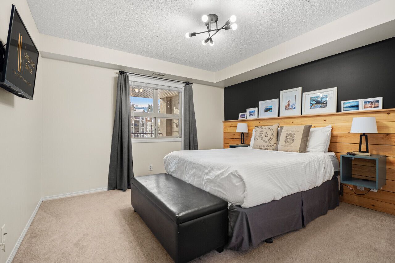A bedroom with a large bed, a bench, a window with curtains, a wall-mounted TV, and framed pictures.
