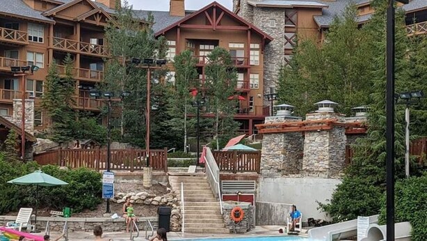 A pool area with a stone wall, wooden deck, and a view of a lodge building. - Nature`s Nook | Ski In/Out | Pools | Hot Tub (Panorama)
