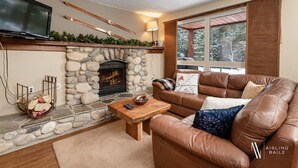 A cozy living room with a stone fireplace, leather sofa, and a television mounted on the wall. - Nature`s Nook | Ski In/Out | Pools | Hot Tub (Panorama)