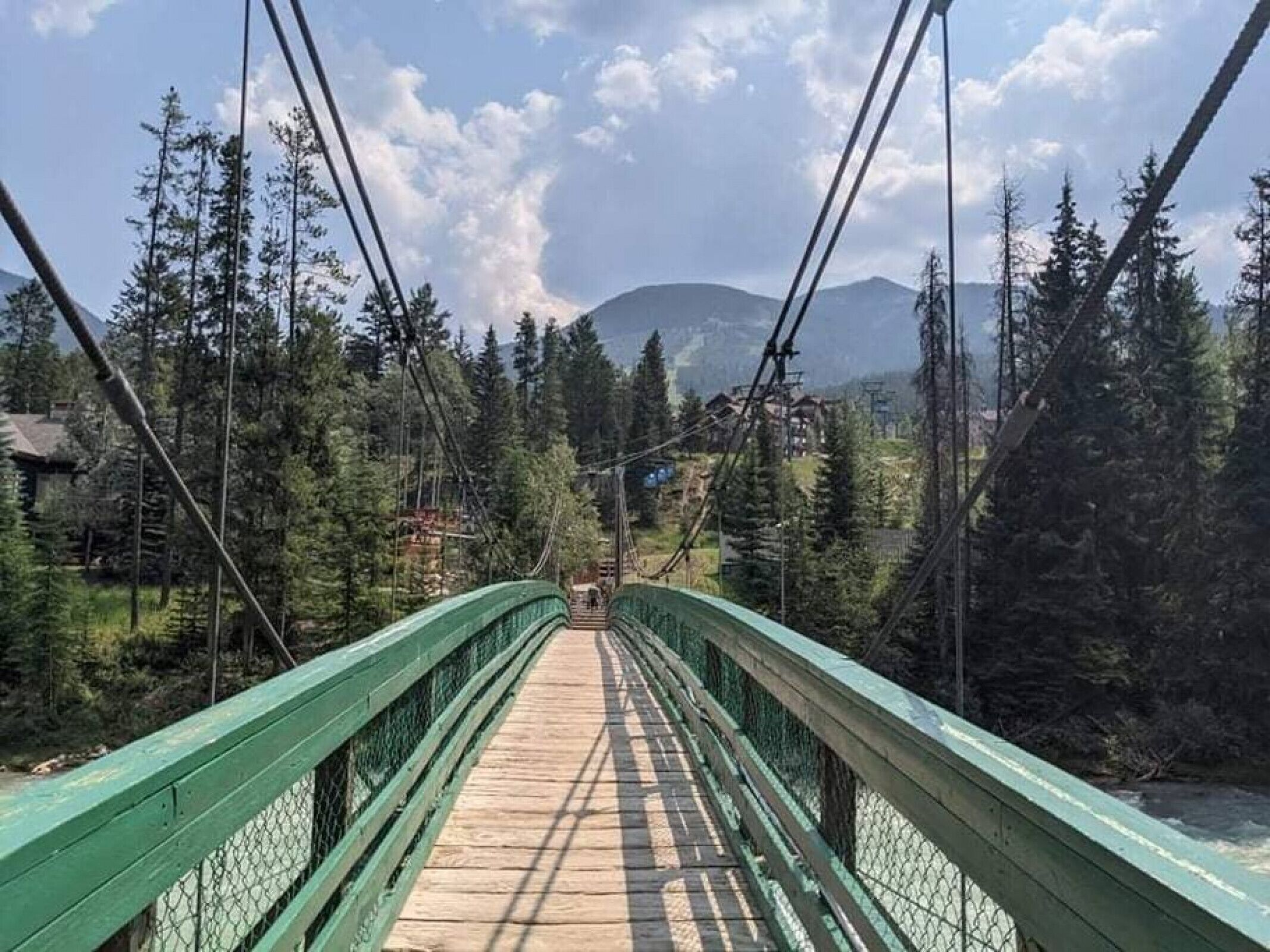 Un puente peatonal de madera con barandillas verdes, rodeado de un bosque denso y una montaña al fondo.