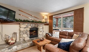 A cozy living room with a stone fireplace, leather sofa, and a television mounted on the wall. - Nature`s Nook | Ski In/Out | Pools | Hot Tub (Panorama)