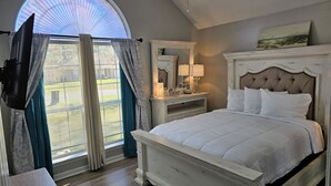 A bedroom with a large bed, a dresser, a mirror, and a TV mounted on the wall. - Beautiful Warm Beach House Retreat, King bed, fireplace and Pool (Shreveport)