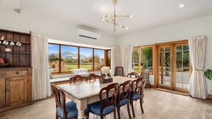 A dining area with a wooden table and chairs, a chandelier, and large windows offering a view of the outdoors.
