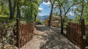 Un chemin de gravier mène à un bâtiment en pierre surmonté d’un toit en tuiles, entouré d’arbres et d’un mur de pierre.