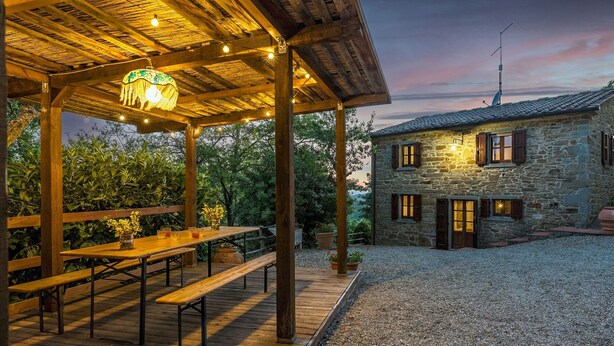 Un bâtiment en pierre avec des bancs et des tables en bois, situés sous une zone abritée et illuminée par des guirlandes lumineuses.