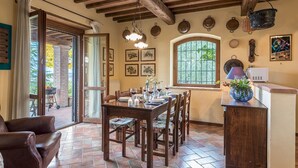 Zona pranzo con tavolo in legno apparecchiato per sei persone, circondato da sedie. Si vede una finestra con vista su piante e, alla destra, un angolo cottura con un forno a microonde e un vaso di fiori.