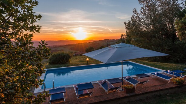 Una piscina con lettini e un ombrellone, immersa nel verde con vista sul tramonto.