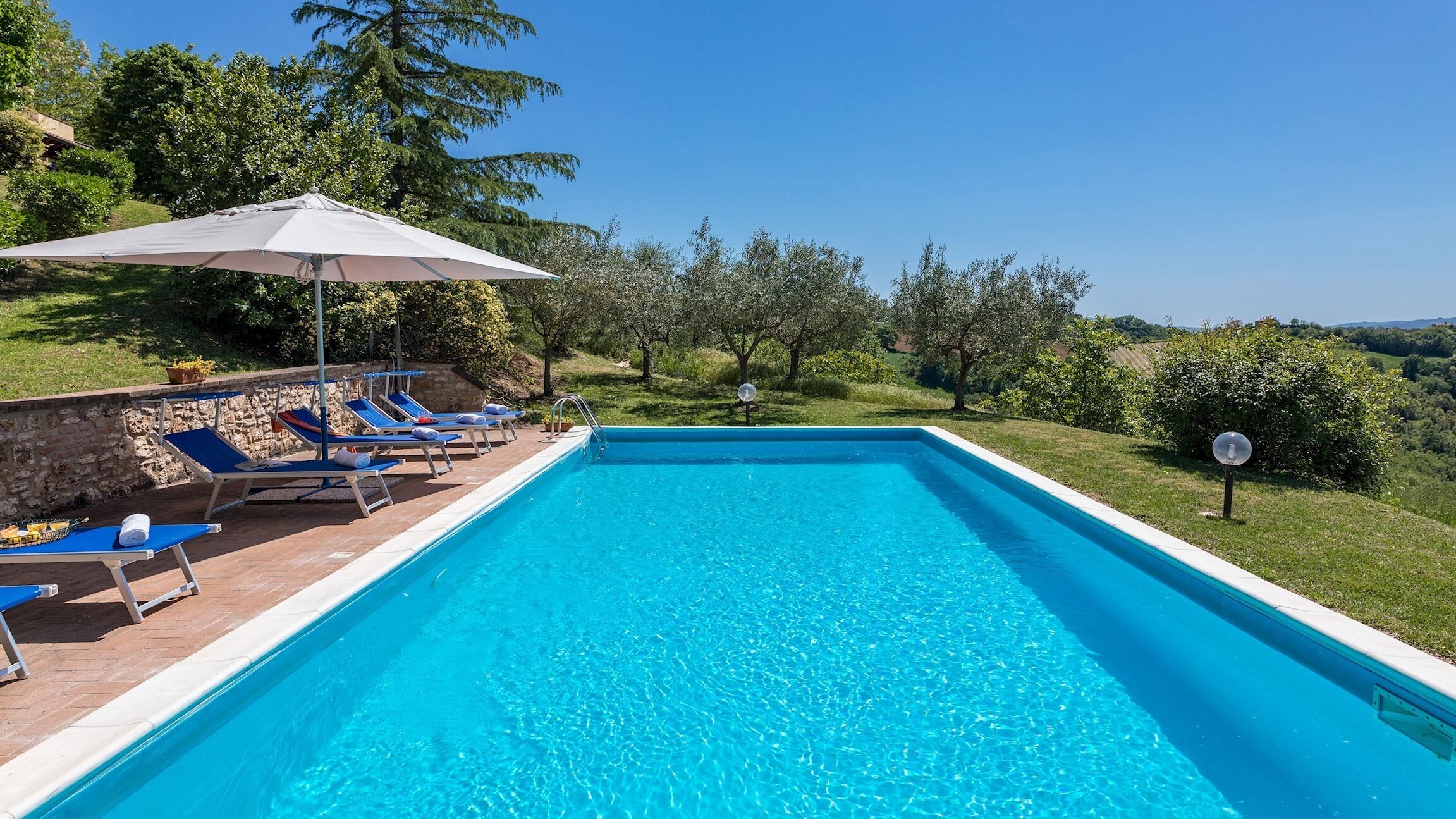 A rectangular swimming pool with clear blue water, surrounded by a stone wall and a row of blue lounge chairs.