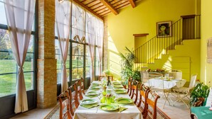 Un espace repas avec une longue table dressée pour le repas, de grandes fenêtres avec des rideaux, un escalier et une vue sur l’extérieur.