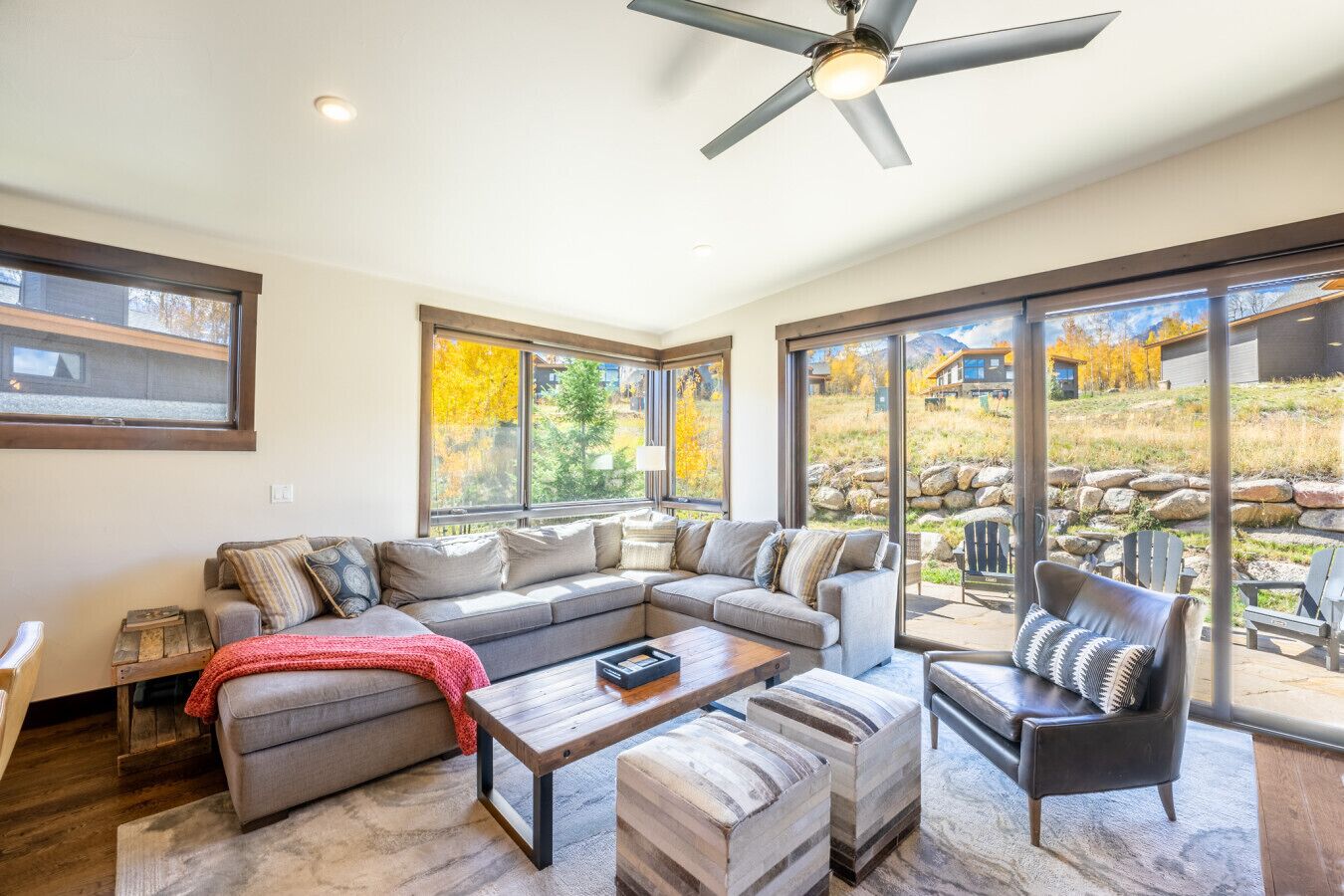 A spacious living room with a sectional sofa, a coffee table, and a fireplace. Large windows offer views of the outdoors.