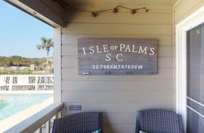 A wooden sign with the text 'Isle of Palms S.C.' and a phone number, mounted on a building with a view of a pool and palm trees.