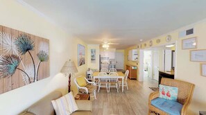 A spacious living area with a dining table, chairs, and a sofa. There is a kitchen in the background. - 2 bed/2bath condo with pool, 2 min walk to beach! (Isle of Palms)