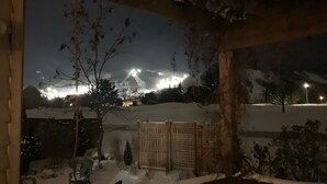Ein verschneiter Außenbereich mit Holzzaun und Blick auf ein hell erleuchtetes Gebäude in der Ferne.
