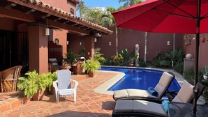 Un espace piscine avec des chaises longues et un parasol rouge, entouré de plantes en pot et d’une terrasse carrelée.