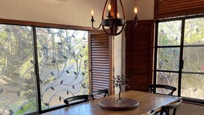 Un espace repas comprenant une table et des chaises en bois, un lustre et de grandes fenêtres donnant sur un paysage verdoyant.
