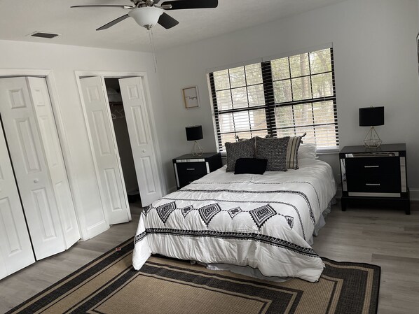 A bedroom with a bed, bedside tables, a ceiling fan, a window with blinds, and a door.